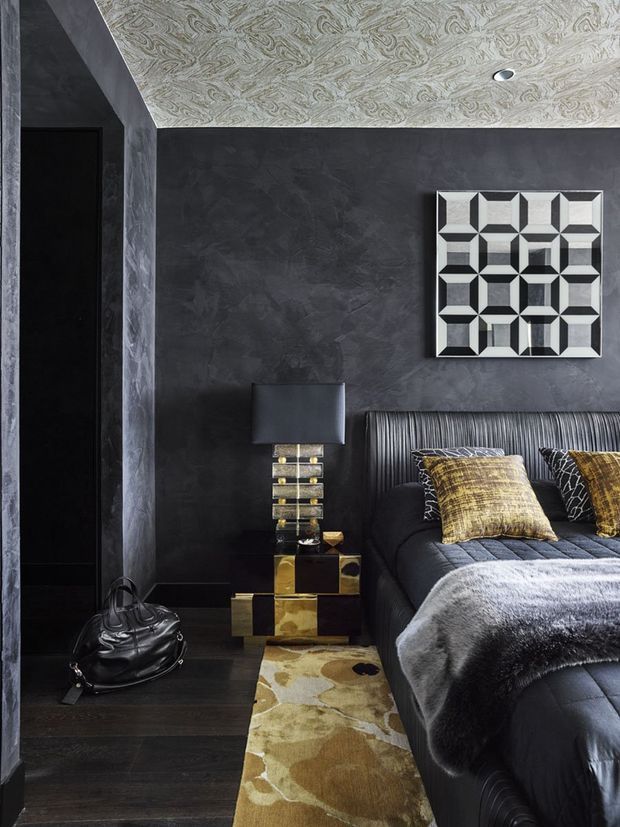 A dark, glamorous bedroom with charcoal textured walls, a patterned light ceiling, and dark wood flooring. The room features a black upholstered bed with a vertically channelled headboard, layered grey and metallic-gold cushions, a plush grey throw, a gold-and-black bedside table, a sculptural table lamp, and a large geometric wall clock. A cowhide-style runner adds contrast beside the bed.