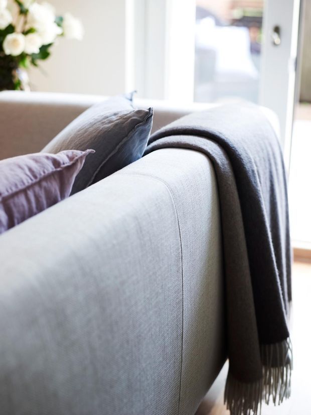 Close-up interior detail of a pale upholstered sofa with layered grey and muted lavender cushions and a soft charcoal throw draped over the back. Bright daylight enters through a glazed door or window, with blurred white floral decor in the background. The image has a soft, calm, neutral styling.