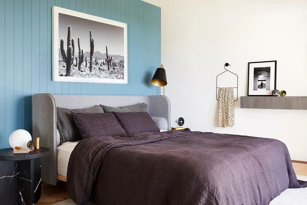 A stylish contemporary bedroom with a grey upholstered bed, dark mauve-purple bedding and cushions, a blue vertical-panel feature wall, and a white adjacent wall. A black-and-brass wall lamp, framed monochrome cactus artwork, floating shelf, framed print, and hanging garment add decorative detail. Timber flooring is visible around the bed.