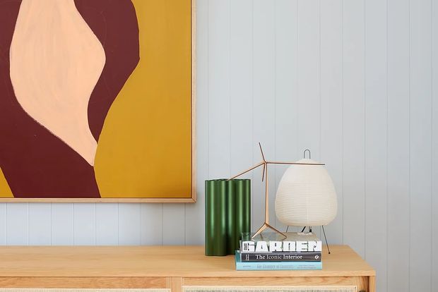 A styled interior vignette featuring a light timber console against pale vertical wall panelling. The display includes a large abstract artwork in a timber frame, green cylindrical decorative vessels, a sculptural white table lamp, and stacked design books.