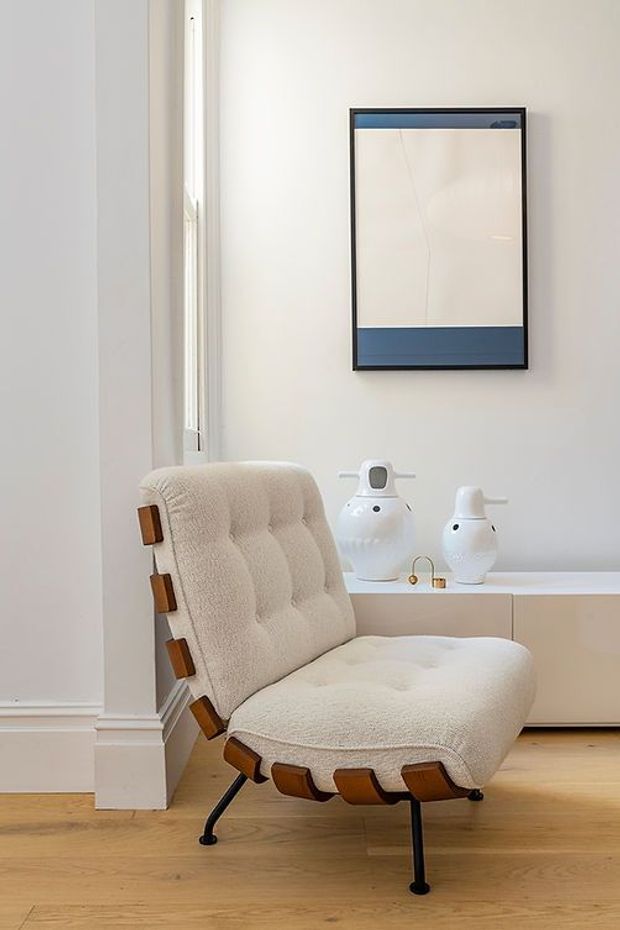 A softly styled interior corner featuring a sculptural cream upholstered lounge chair with visible wooden side supports and dark metal legs. The chair sits on a light timber floor beside a low white built-in cabinet, with two white decorative vases and abstract blue-and-white wall art. White walls, classical skirting and trim, and tall narrow glazing create a bright, refined setting.
