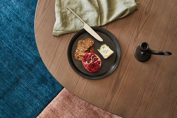 Overhead view of a round warm wood coffee table with a dark ceramic plate holding bread, cheese and pomegranate, alongside a small dark vessel and folded linen napkin. The table sits over layered blue and blush textured rugs, creating a warm, tactile interior styling scene.