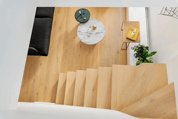 Top-down view of a light timber staircase descending into an open-plan living area. The space features matching timber flooring, a dark upholstered sofa, round coffee tables, a slim wall-mounted side table, and a potted plant. The staircase has broad pale-wood treads and a distinctive angled landing, with clean white walls and a minimalist contemporary feel.
