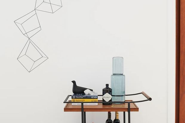 A minimalist interior vignette featuring a slim mid-century-style timber console with black metal framing and handles, styled with stacked books, a sculptural black bird, bottles, and two pale blue-green glass vessels. The console sits against a plain white wall with geometric wire artwork, beside a warm timber door or window frame.