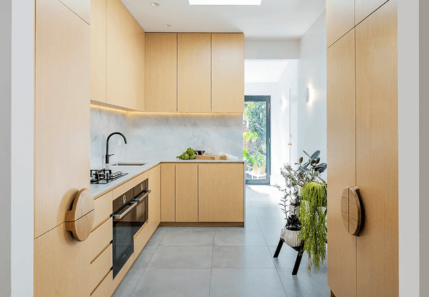 A compact, contemporary galley kitchen with pale timber cabinetry, integrated appliance fronts, a pale stone or marble-look splashback, and large grey floor tiles. The kitchen opens toward a bright hallway or entry area with indoor plants and a glazed door at the rear. Recessed ceiling lighting and under-cabinet illumination create a clean, warm, minimalist feel.