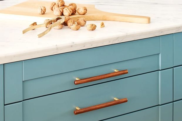 Close-up of a blue kitchen island or cabinetry with shaker-style drawer fronts and long warm wood handles with brass-toned mounts. A white marble-look countertop sits above, with a wooden cutting board and walnuts arranged on the surface.