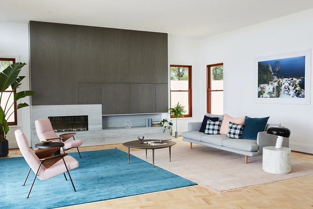 Bright contemporary living room with a large grey textured feature wall and fireplace, pale timber flooring, expansive white walls, and warm timber-framed windows. The room is furnished with a light grey sofa, pink lounge chairs, a low oval coffee table, a blue area rug, and a small cylindrical side table. A framed artwork and indoor plant add restrained decorative accents.