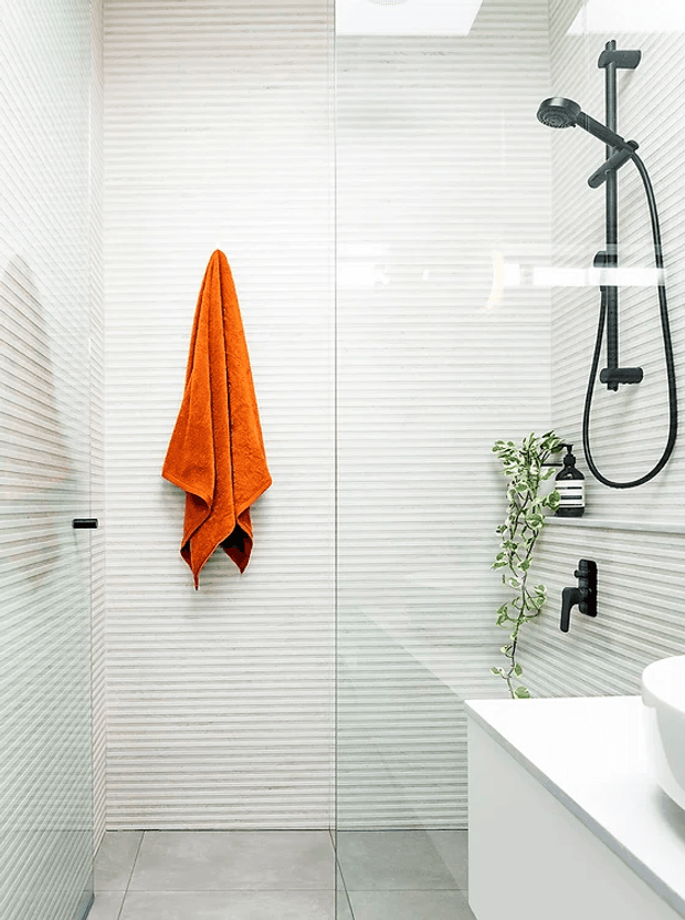 Bright contemporary bathroom shower area with a frameless glass enclosure, horizontally textured light wall tiles, large grey floor tiles, matte black shower fittings, and a white vanity with integrated basin. An orange towel and trailing green plant add contrasting colour.