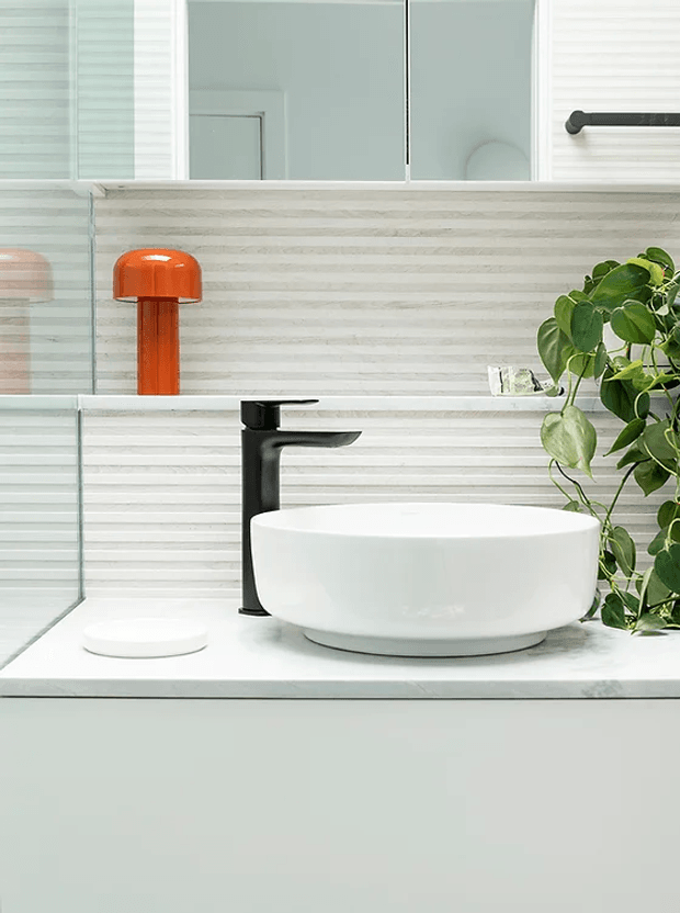 A contemporary bathroom vanity vignette featuring a white vessel basin on a pale stone or solid-surface countertop, paired with a matte black tap. The wall is finished with horizontal light grey and white textured tiles, with mirrored cabinetry above. A bright orange table lamp and leafy green plant add colour accents, while a glass shower screen is visible at the left.