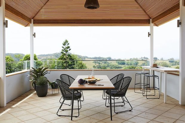 Bright, open-air covered dining terrace with a timber slatted pitched ceiling, white structural posts, glazed balustrades, tiled flooring, a dining table with woven chairs, and a compact bar ledge. The space overlooks a green rural landscape and is furnished in a relaxed contemporary style.