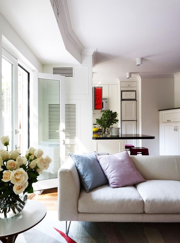 Bright, refined open-plan living room with a light upholstered sofa in the foreground, pastel blue and blush cushions, a round side table with flowers, and a compact kitchen visible behind. The room features white built-in cabinetry, panelled doors, louvred ventilation panels, decorative cornices, timber flooring, large glazed doors, and a dark kitchen island benchtop.