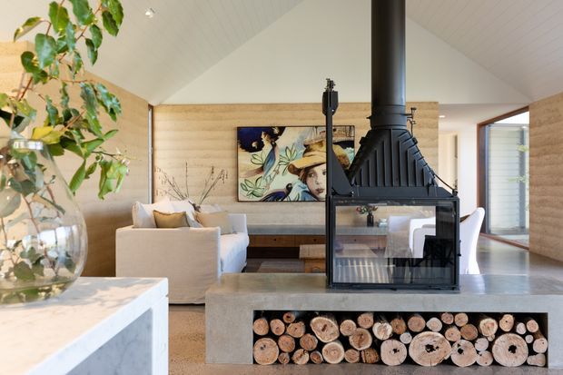 Bright contemporary living room with a pitched white ceiling, pale timber wall panelling, a central black wood-burning fireplace, and a concrete hearth with integrated firewood storage. A light sofa seating area faces an artwork feature wall, while large openings connect the room to adjacent spaces and the outdoors.