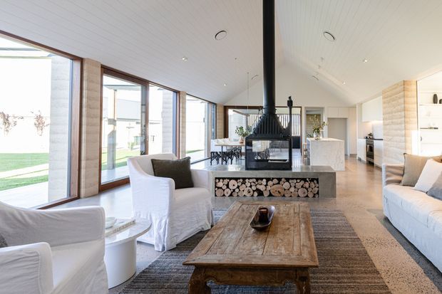 Bright open-plan living area with a freestanding black wood-burning fireplace and log storage feature, white slipcovered seating, a rustic timber coffee table, and large timber-framed windows. The space opens into a contemporary kitchen and dining area, with polished concrete flooring and light timber wall detailing.