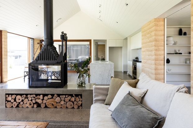 Bright open-plan living area with a large upholstered sofa in the foreground, a central freestanding black wood-burning fireplace with integrated log storage, and a kitchen beyond. The space has a white-lined vaulted ceiling, pale built-in cabinetry and open shelving, a light stone kitchen island, timber-framed glazing, and a warm natural material palette.