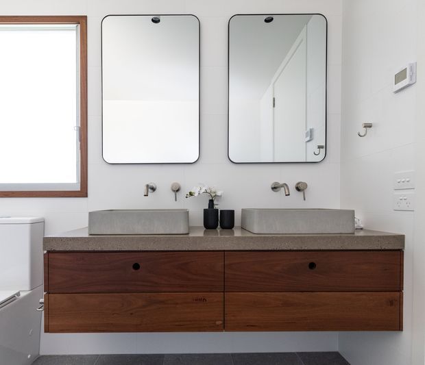 Bright contemporary bathroom featuring a floating double vanity with rich dark timber drawers, a speckled stone benchtop, two rectangular concrete-look vessel basins, wall-mounted brushed metal tapware, and matching rounded-corner mirrors. White walls, a timber-framed window, and dark floor tiles complete the clean, understated fit-out.