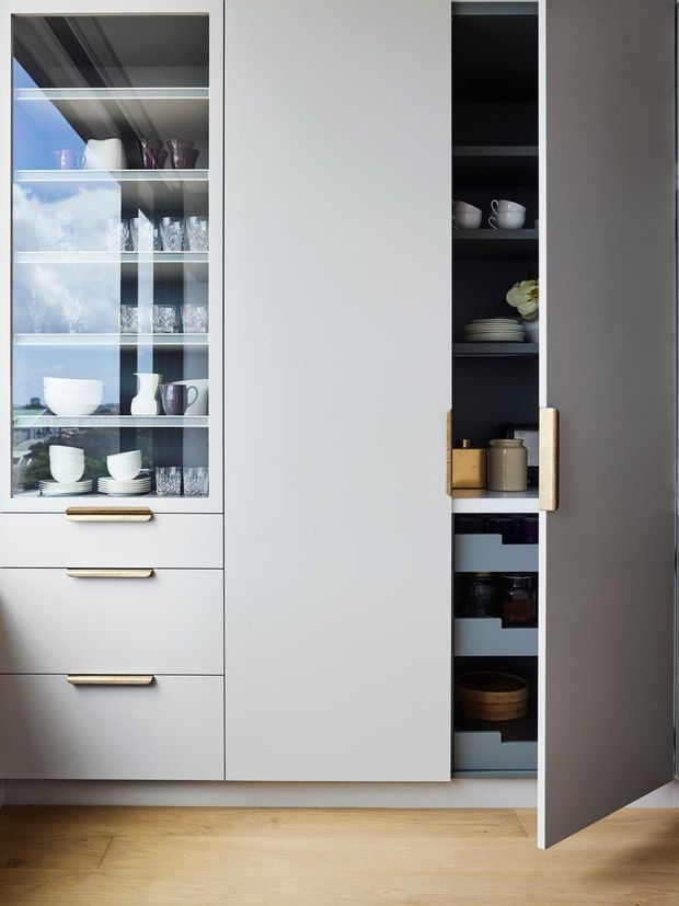 Close-up view of a contemporary kitchen storage wall featuring pale grey full-height cabinetry, a glass-front display cabinet with crockery and glassware, brass-toned drawer pulls, and a narrow open pantry cupboard concealed behind a tall door. The cabinetry stands over a light timber floor and uses dark interior shelving for contrast.