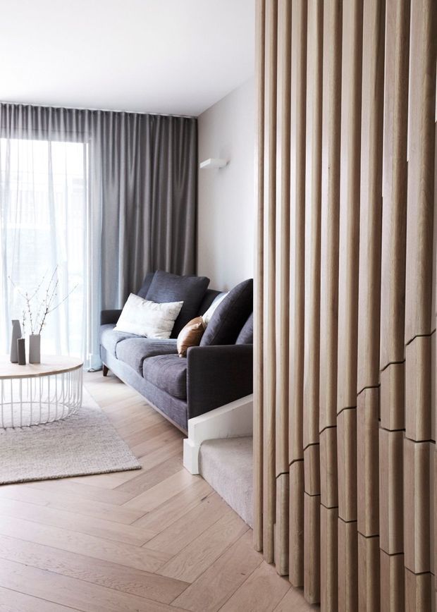 Bright contemporary living room with a dark grey sofa, sheer and charcoal curtains, pale herringbone timber flooring, a round coffee table, and a sculptural vertical timber slat feature wall in the foreground. A short carpeted stair is visible beside the sofa. The space has a calm, refined neutral palette with strong natural wood detailing.