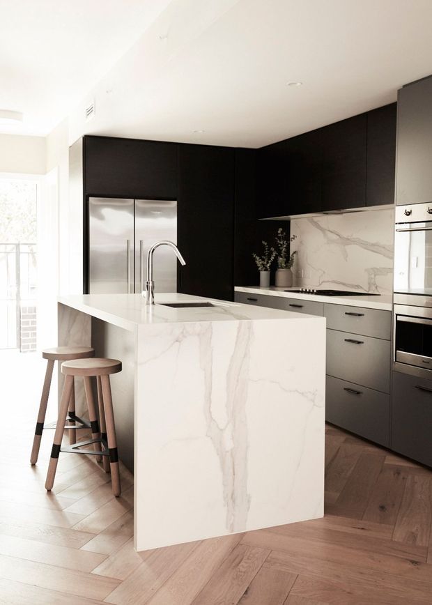 Contemporary kitchen featuring a large white marble-look island with waterfall edge, dark matte cabinetry, integrated stainless-steel appliances, and a marble-look splashback. Two timber stools sit at the island, while warm herringbone timber flooring and abundant natural light soften the monochrome palette.