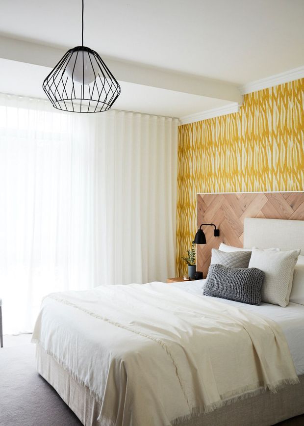 Bright, contemporary bedroom featuring a neatly made upholstered bed with layered white and grey bedding, a timber herringbone headboard, bold mustard-yellow patterned wallpaper, sheer white curtains, and a black geometric pendant light. The room has a soft, warm, refined atmosphere with strong natural light.