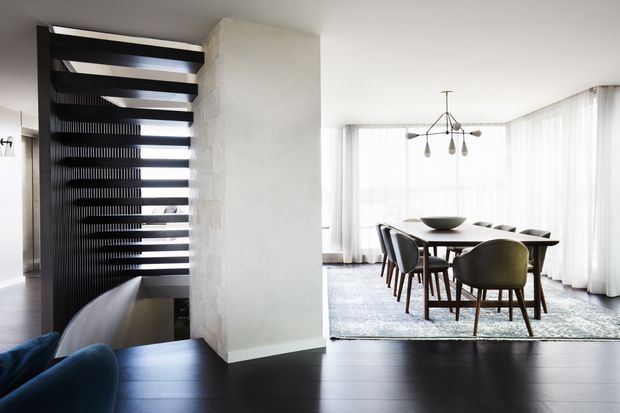A refined open-plan dining area viewed from an adjacent living space, featuring a dark timber floor, a long dining table with upholstered chairs, sheer floor-to-ceiling curtains, and a sculptural pendant light. A dramatic black open-riser stair screen with horizontal treads and vertical slats forms a strong architectural feature beside a pale textured wall.
