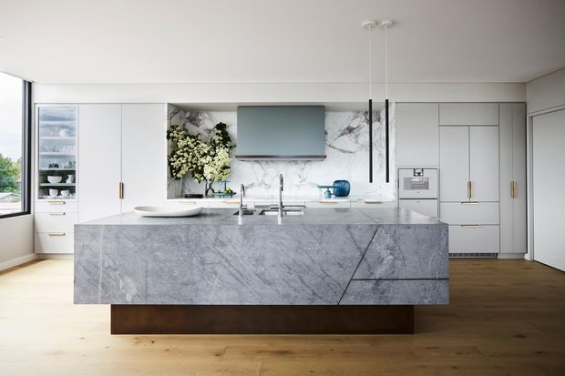 Contemporary open-plan kitchen featuring a large waterfall-edge grey-veined stone island with integrated cooktop and sink, white full-height cabinetry, marble-look backsplash, timber flooring, and a concealed rangehood beneath a wall-mounted screen or appliance. The space is bright and refined, with pendant lighting and subtle brass hardware.