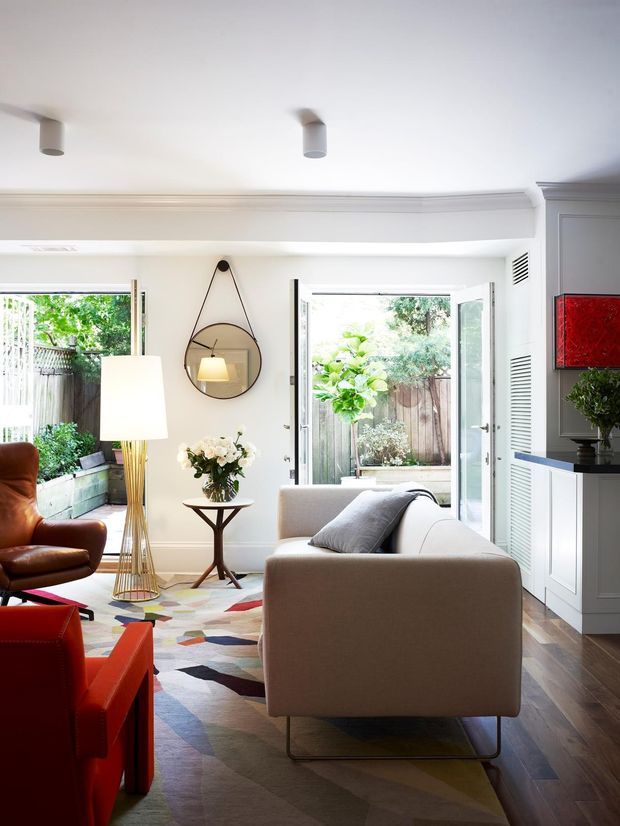 Bright, refined living room with white walls and decorative ceiling cornice, timber flooring, a large geometric multicolour rug, upholstered sofa, leather lounge chairs, sculptural floor lamp, side table and floral arrangement. Full-height glazed doors open to a small planted courtyard or garden, while built-in white cabinetry and a red artwork add visual interest.