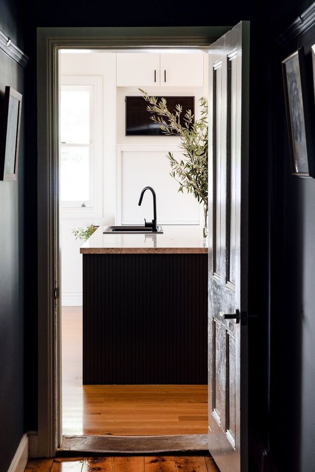 View through a dark, paneled doorway into a bright compact kitchen. The kitchen features white cabinetry, a black vertical-groove island front, a warm timber benchtop, black gooseneck tap and sink, timber flooring, and a wall-mounted television or dark display panel. A tall leafy branch adds a natural accent beside the island.