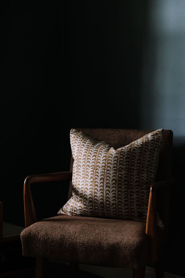 A close-up, moody interior photograph of a dark brown upholstered armchair with wooden arms and legs, positioned against a nearly black wall. A patterned beige and brown decorative cushion rests on the chair, with dramatic side lighting creating strong shadows and a refined, intimate atmosphere.