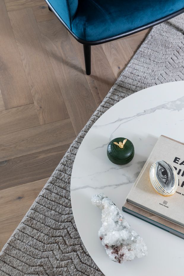 Close-up interior vignette showing a white veined oval coffee table on a textured grey rug, pale oak flooring, and the edge of a deep teal upholstered chair. Decorative objects, books, and a crystal-like ornament are arranged on the table.