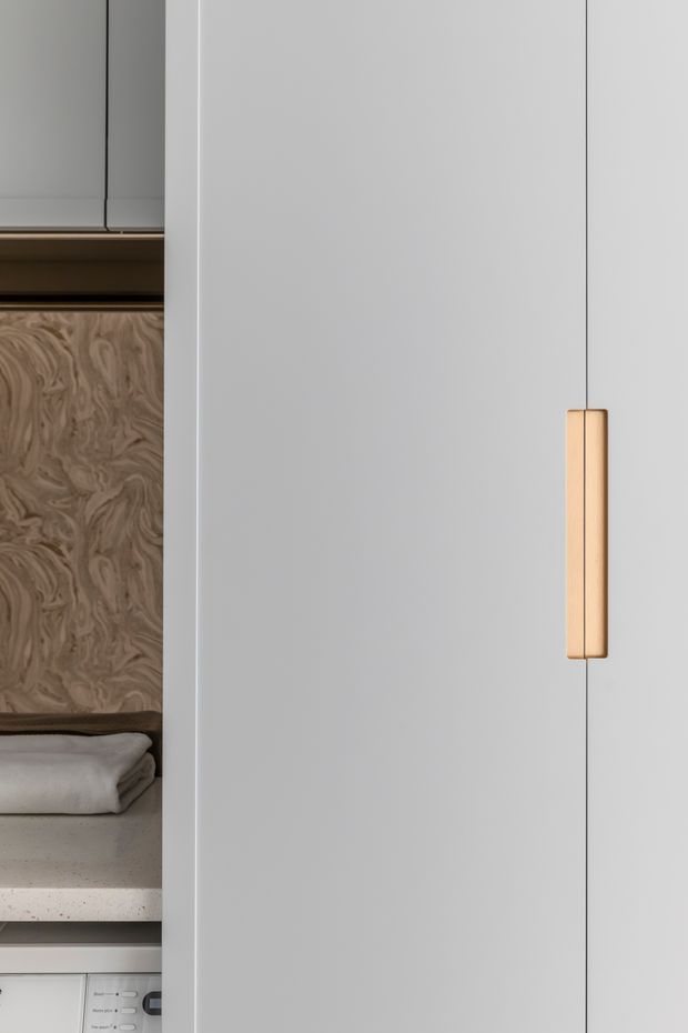 Close-up view of a minimalist built-in white cabinetry wall, featuring a slim warm timber or brass-toned vertical handle. An open compartment at left reveals a swirling wood-grain feature panel, folded textiles, a pale stone-like shelf, and part of a built-in appliance below.