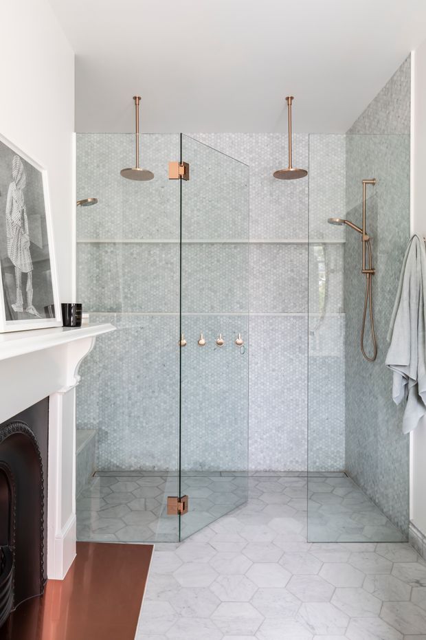 Bright, refined bathroom featuring a large frameless glass walk-in shower enclosed with bronze-toned hardware. The shower walls are clad in pale blue-grey small-format mosaic tile, while the floor uses large white and light-grey hexagonal tiles. A classic white fireplace surround is visible at left, with a framed artwork, wall-mounted shower fittings, a towel rail, and soft white towels.