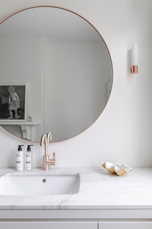 Bright, refined bathroom vanity with a large round mirror edged in rose-gold metal, a white inset basin, pale marble countertop, white cabinetry, and a coordinated wall sconce and faucet in a warm metallic finish. The styling is clean and contemporary with subtle classic detailing, including a reflected mantel-like shelf and framed artwork.