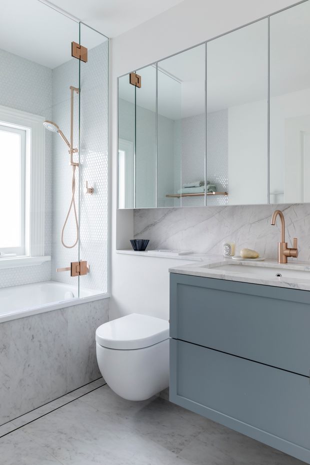 Bright, compact contemporary bathroom featuring a freestanding-style bathtub with glass shower screen, wall-mounted toilet, mirrored wall cabinetry, and a muted blue-grey vanity. Pale marble-look wall and floor surfaces are paired with white hexagonal shower tiles and warm brushed copper fixtures.