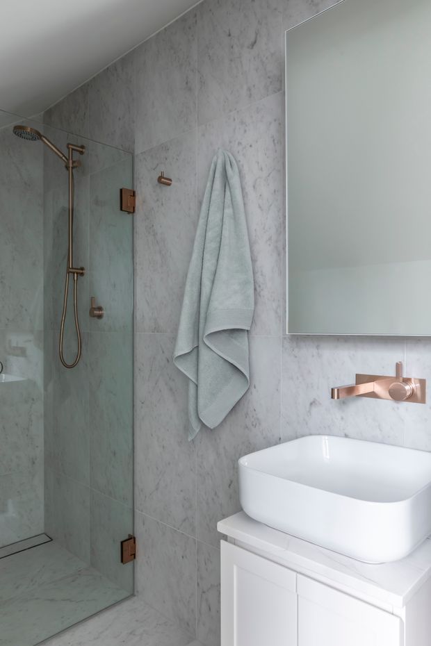 A compact, refined bathroom with full-height pale grey marble-look wall and floor tiles, a frameless glass shower with rose-gold/copper fixtures, a wall-mounted towel hook, a large frameless mirror, and a white vessel basin on white built-in cabinetry. The restrained palette and metallic accents create a clean, contemporary feel.