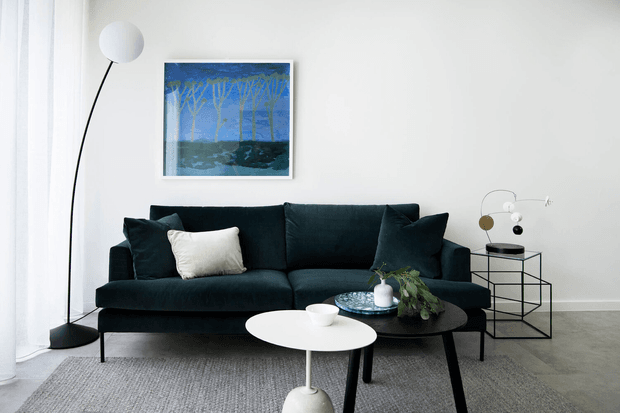 A refined contemporary living room with a deep charcoal sectional sofa, black accent cushions, a white coffee table, a dark side table, and a sculptural arc floor lamp. A framed blue-toned artwork hangs above the sofa, with a geometric black metal console and decorative objects to the right. The room has white walls, sheer curtains, a grey area rug, and a restrained monochromatic palette with blue and greenery accents.