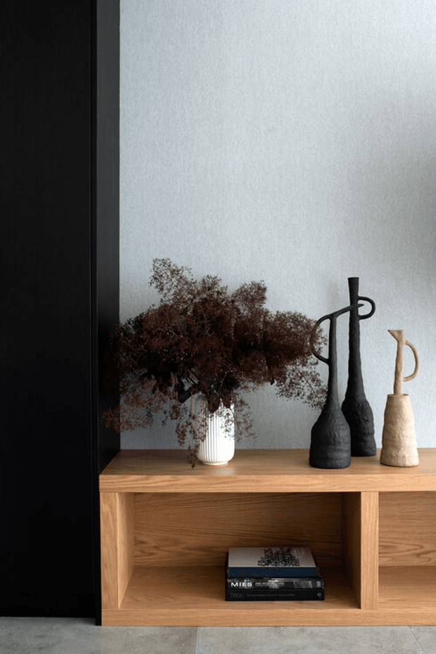 Close-up interior vignette of a warm timber open shelving console against a pale textured wall. The shelf displays sculptural black and beige ceramic vessels, a white ribbed vase with dried foliage, and stacked books. A dark vertical panel frames the left side, while a grey floor is visible below.