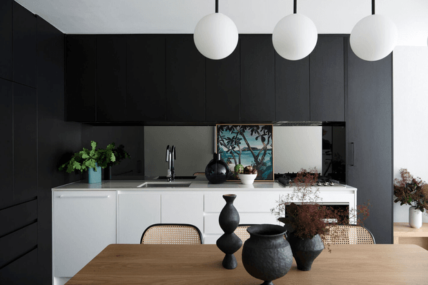 Contemporary open-plan kitchen and dining area with a long white island, integrated dark cabinetry, timber dining table, cane-backed chairs, sculptural black vases, and three spherical pendant lights. A framed artwork and indoor plants add restrained natural accents against the monochrome palette.