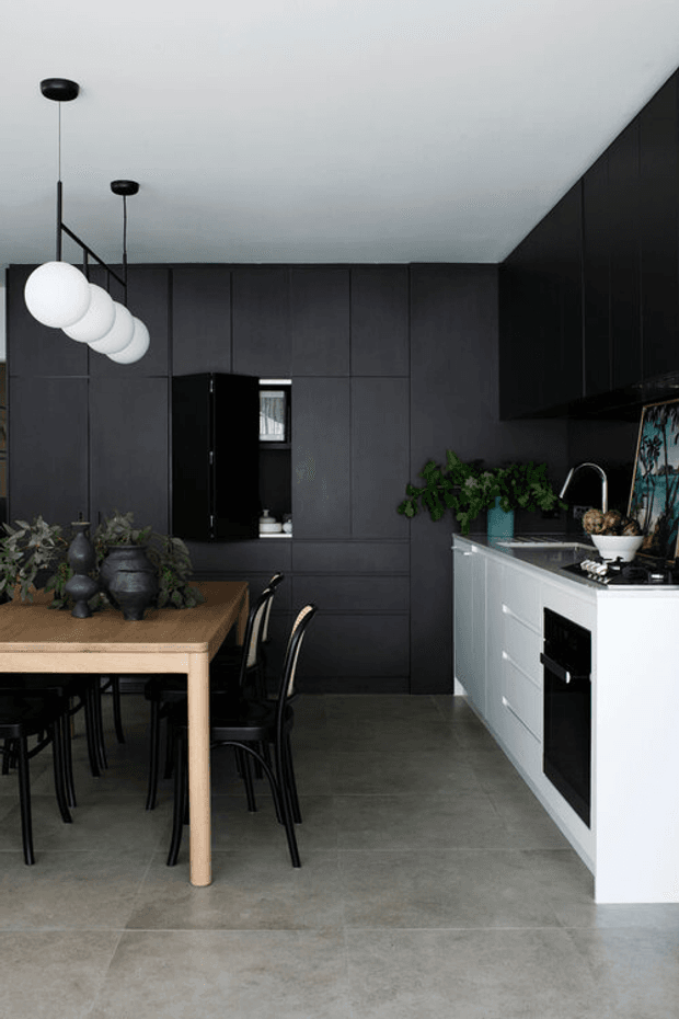 A contemporary kitchen and dining area with dark charcoal full-height cabinetry and walls, a white island with integrated black appliances, grey tiled flooring, and a timber dining table with black chairs. Minimal black pendant lights hang above the dining area, while plants and decorative objects add organic contrast.
