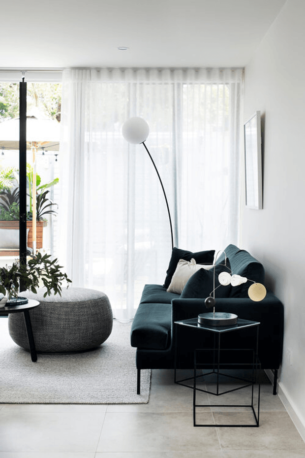 Bright contemporary living room with a dark teal sectional sofa, sculptural black arc floor lamp, nesting side tables, large round upholstered ottoman, sheer floor-to-ceiling curtains, a light textured rug, and pale large-format floor tiles. Indoor planting and a glazed opening connect the room to an outdoor courtyard or garden.