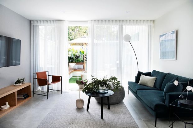 Bright contemporary living room with a deep teal sectional sofa, black round coffee table, timber media console, and a pair of rust-toned lounge chairs. Sheer floor-to-ceiling curtains provide abundant filtered daylight, with indoor plants, pale flooring, and minimalist artwork creating an airy, refined setting. A small outdoor courtyard or balcony is visible through the glazing.