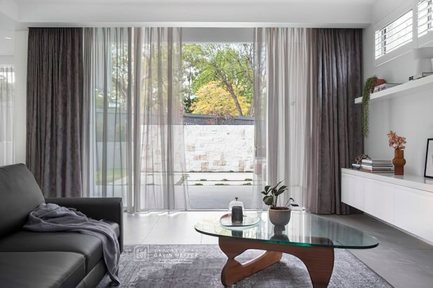 A bright, refined living room with floor-to-ceiling glazed doors framed by layered sheer and dark drapery, opening to a small paved courtyard. The room features a grey sectional sofa, glass coffee table with sculptural timber base, pale grey rug, white built-in media cabinetry, display shelving, and high clerestory-style windows.