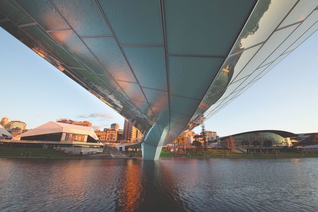 A wide-angle exterior architectural photograph looking along the underside of a large sculptural pedestrian bridge spanning a river. The bridge has a sweeping, cantilevered form with reflective and paneled metal soffit surfaces, supported by a central sculptural pier. An urban riverside setting with mid-rise buildings and a modern curved-roof building is visible beyond the water.