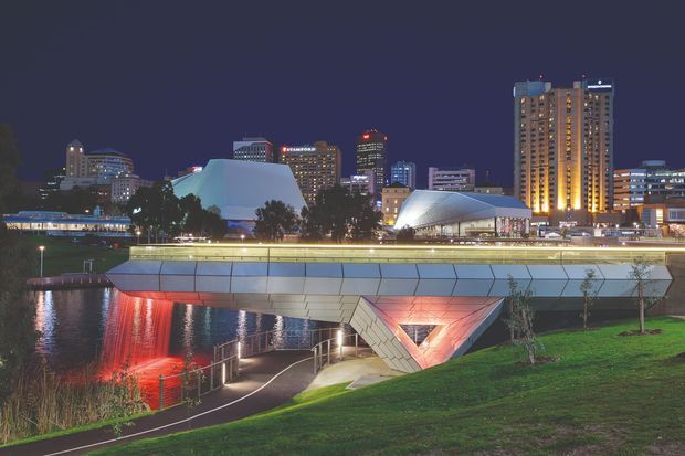 Night-time exterior view of a modern urban waterfront precinct, showing a broad angular pedestrian bridge or elevated public structure over a waterway. The structure has faceted light-grey cladding, illuminated linear handrails, and a dramatic triangular support or undercroft glowing red from architectural lighting. A landscaped grassy bank and riverside walkway occupy the foreground, with illuminated contemporary civic and high-rise buildings forming the background skyline.
