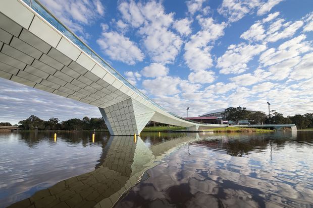 A wide exterior photograph of a striking modern pedestrian bridge spanning a calm body of water. The bridge has a sweeping, sculptural form with a pale tiled or panelled underside, angled support pier, slim glass or metal balustrades, and strong reflections in the water. A low modern building and landscaped shoreline sit in the background beneath a partly cloudy sky.