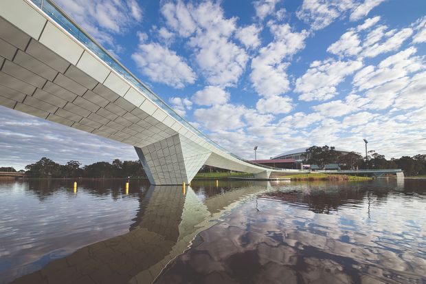 Exterior architectural photograph of a long, sweeping pedestrian bridge spanning calm water. The bridge has a dramatic cantilevered form with a faceted pale metal-clad soffit and angular support, reflected in the water beneath. A low modern building and landscaped shoreline appear in the background under a partly cloudy sky.