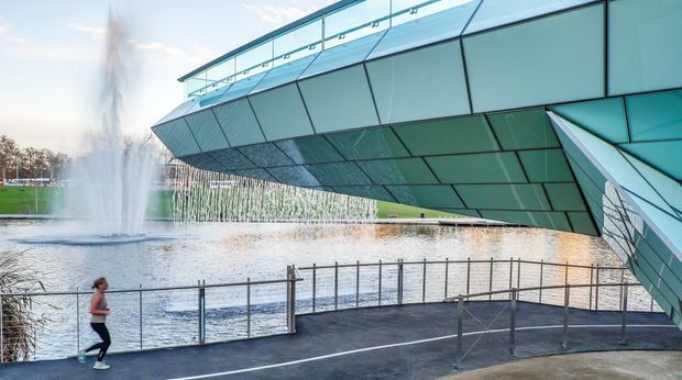 Exterior architectural photograph of a dramatic cantilevered, angular building clad in blue-green glazed panels, projecting over a waterfront promenade. A glass balustrade lines the paved walkway, with a large fountain and water feature beside the structure. The image emphasizes the building envelope, sharp geometric form, extensive glazing, and cantilever.