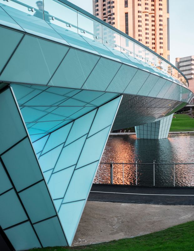 Close-up exterior architectural view of a dramatic contemporary pedestrian bridge or building structure spanning a water feature in an urban setting. The structure has faceted turquoise-blue glazed or metal-clad surfaces, angular supports, a transparent glass balustrade, and strong geometric panel lines. High-rise buildings are visible in the background, with landscaped grass, paving, and reflected light on the water below.