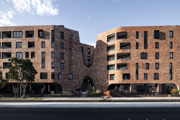 Street-facing view of a contemporary multi-storey apartment building with two curved brick-clad wings connected by a recessed central section. The facade features regularly spaced dark-framed windows, recessed balconies, a prominent arched entrance, and landscaped street-level planting.