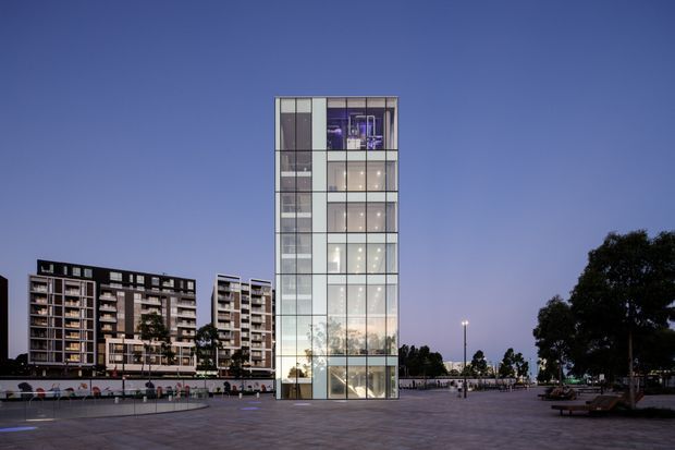 Twilight exterior view of a contemporary multi-storey glass tower with a highly transparent facade, illuminated interior floors, and a broad paved public plaza. Additional apartment buildings and landscaped public-space elements are visible in the background.