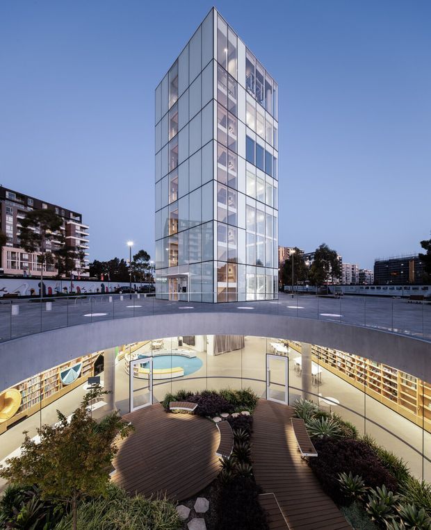 Twilight exterior view of a contemporary multi-storey glass tower rising from a sunken landscaped courtyard. The transparent pavilion-like building has a gridded glazed facade and a sharply pointed roofline, while the lower level contains a brightly lit commercial or library-like interior with extensive shelving, curved circulation areas, timber decking, planting, benches and a circular reflecting or decorative feature.
